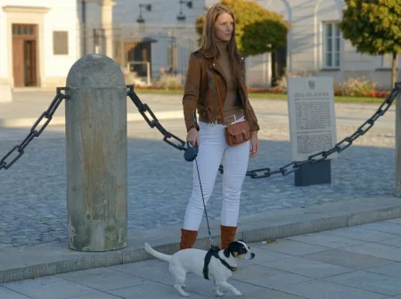 A woman in a brown leather jacket and white jeans walks a small white and black dog on a leash through a cobblestone plaza with historic buildings.