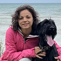 A smiling Ala Krivov wearing a bright pink jacket and posing with Orca, a large black dog with its tongue out, in front of a calm Lake Huron.